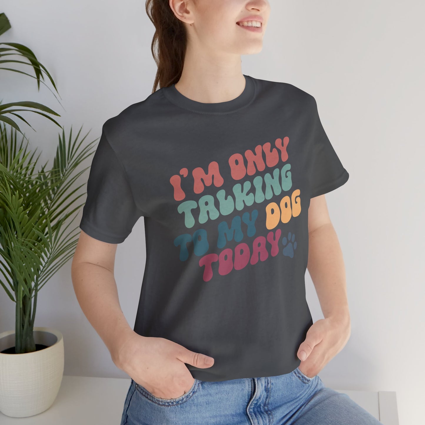 A person grins, donning the Banks + Cove Funny Dog Lover Tee proudly proclaiming, Im only talking to my dog today. Ideal for dog enthusiasts and gifting occasions, they stand with hands in pockets beside a green potted plant.