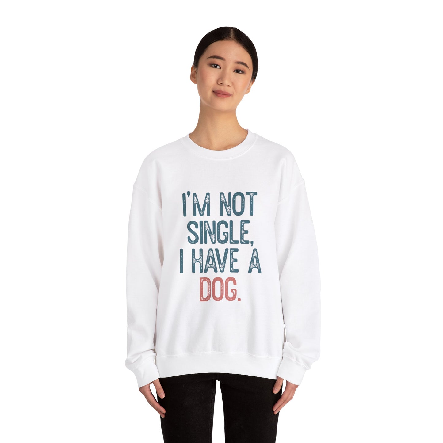 A person with dark hair stands against a white backdrop, wearing the Printify Dog Lover Crewneck Sweatshirt - I’m Not Single. The blue and pink design humorously states IM NOT SINGLE, I HAVE A DOG, making it an ideal statement piece for dog lovers or as a gift for pet owners.