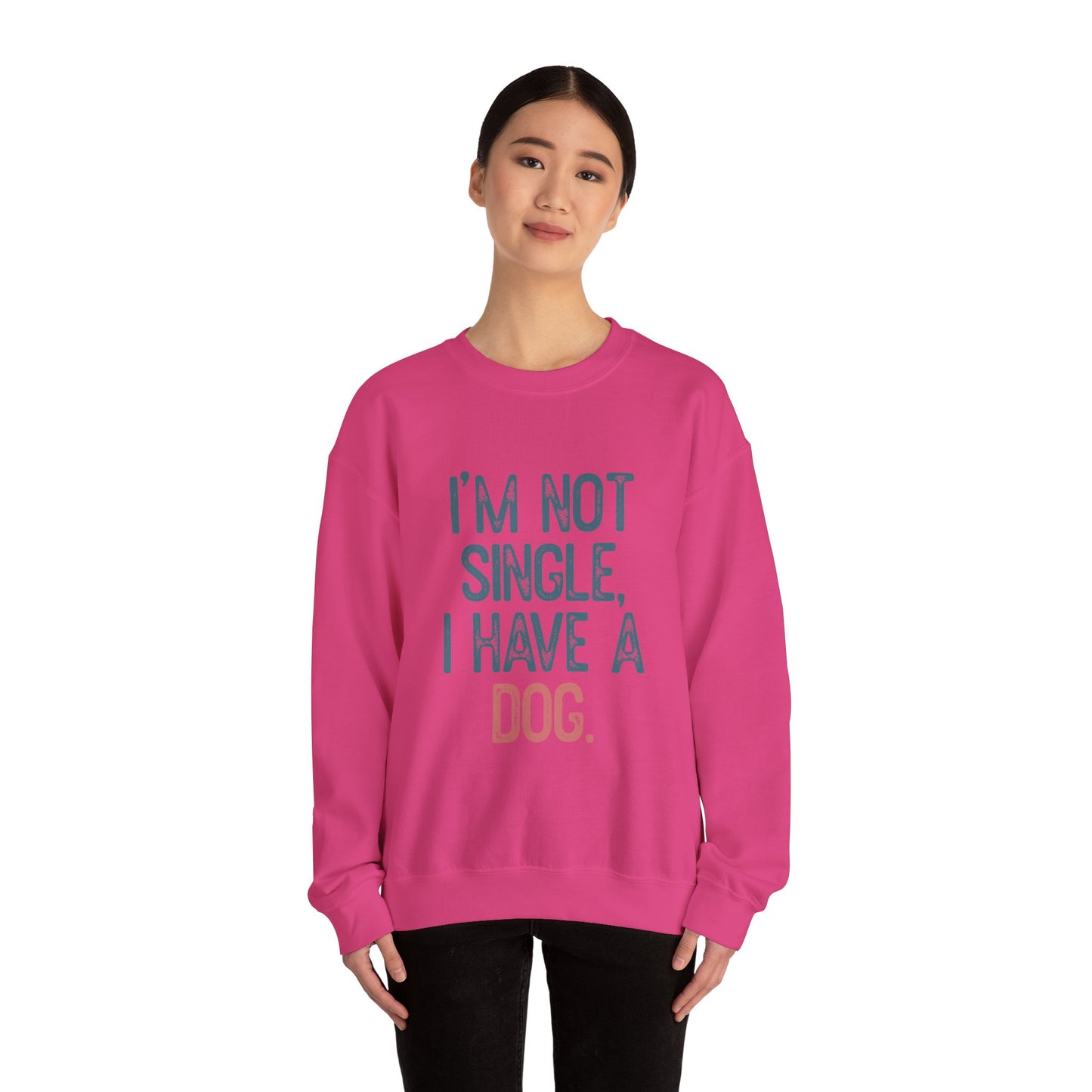 A person with dark hair and black pants wears the Banks + Cove Dog Lover Crewneck Sweatshirt - I’m Not Single, featuring the text Im not single, I have a dog, stands against a plain background, smiling slightly. This pink unisex sweatshirt makes an ideal gift for pet owners.