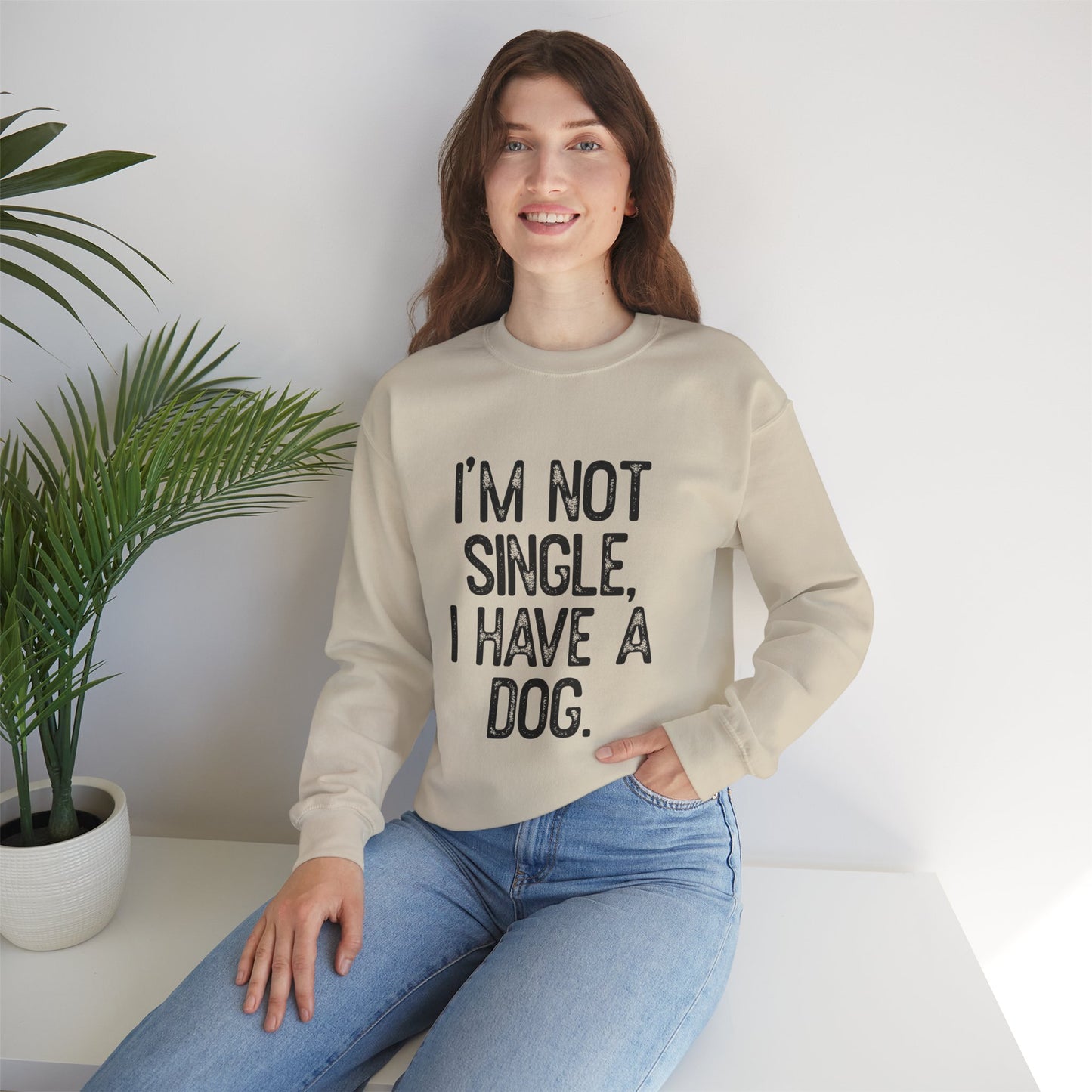 A woman smiles while sitting on a white surface, wearing Printifys Funny Dog Lover Crewneck Sweatshirt with IM NOT SINGLE, I HAVE A DOG text. Styled with light blue jeans and accompanied by a green potted plant, this unisex sweatshirt is ideal for dog lovers who appreciate comfort and style.