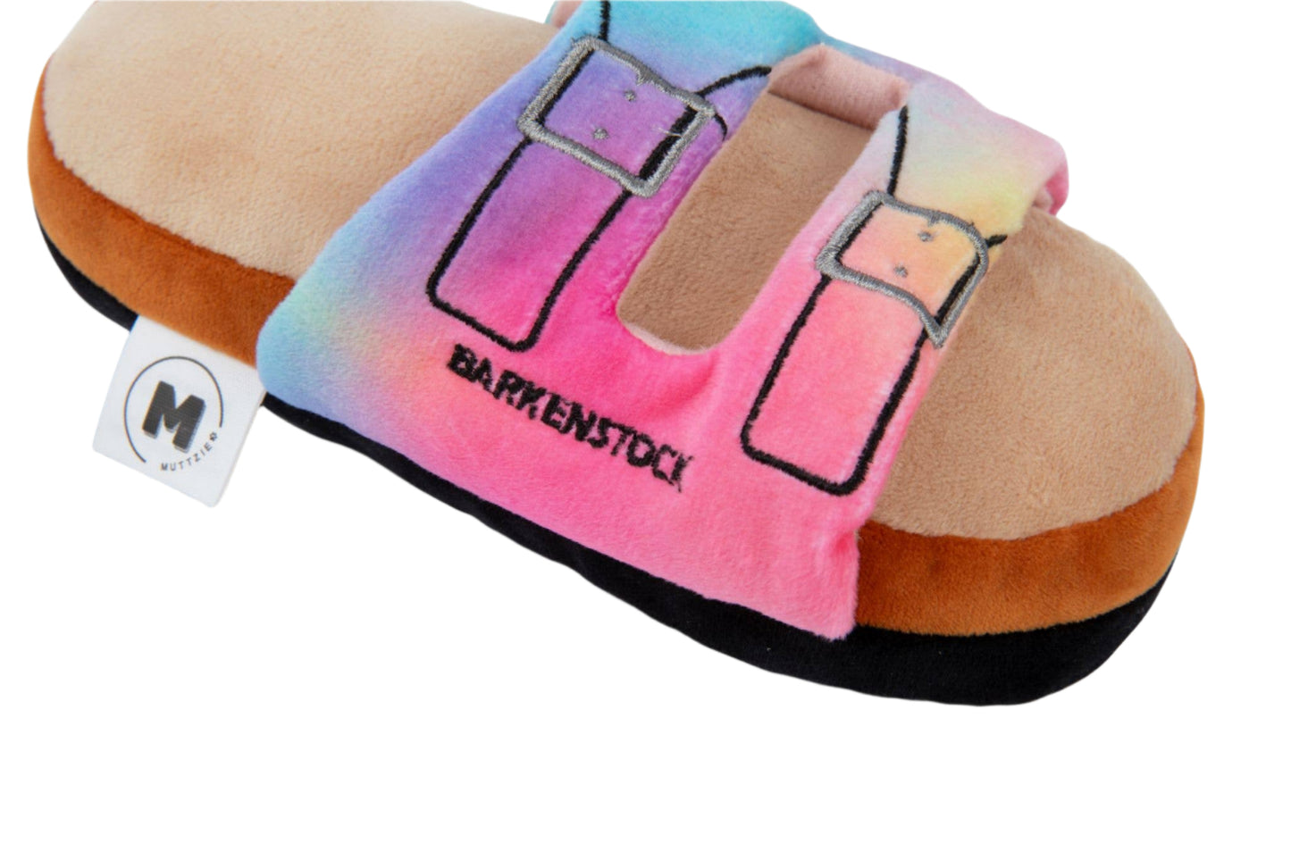 The Barkenstock Rainbow Sandal Toy by Muttzie is a plush delight for dogs, featuring a tie-dye pattern in pink, blue, and purple. It has embroidered straps with Barkynstock text and includes a double squeaker surprise. A black M logo appears on the white tag.
