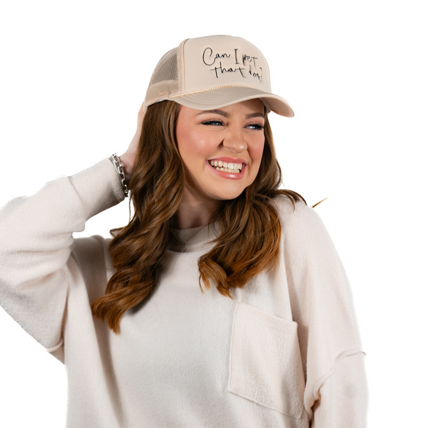 A joyful woman with long wavy hair wears a Banks + Cove Can I Pet That Dog? beige trucker hat and a light-colored sweater, touching the brim of her hat against a plain white background.