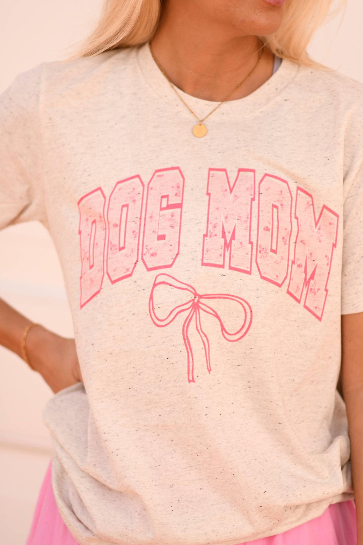 An individual in a gray Dog Mom Bow Tee by P&PD Wholesale, adorned with bold pink letters and an embroidered bow design, enjoys the cozy feel of Bella Canvas fabric. The ensemble enhances their long blond hair and gold necklace.