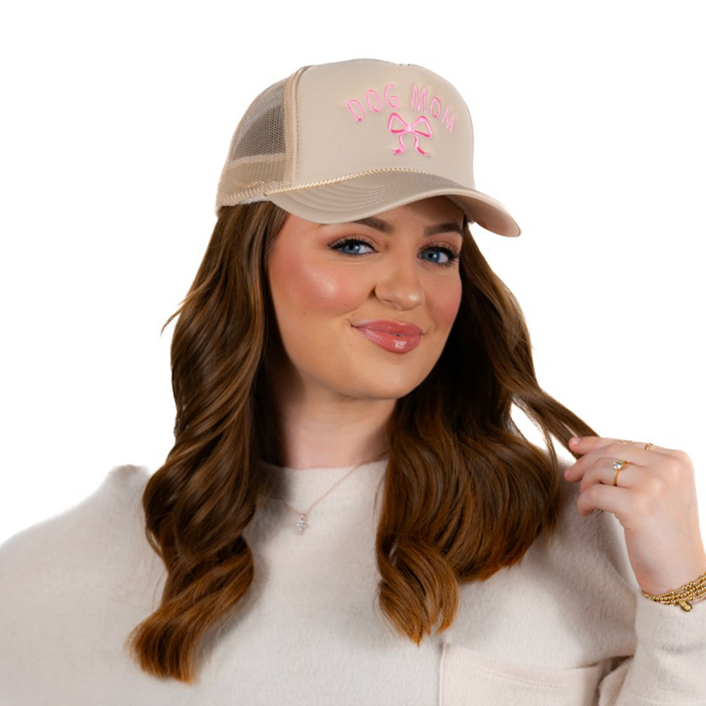 A woman with long brown hair smiles, wearing a beige Banks + Cove Dog Mom Bow Trucker Hat in pink letters. Dressed in a light sweater, she lovingly holds a strand of her hair against a plain white background, celebrating her bond with her four-legged friends.