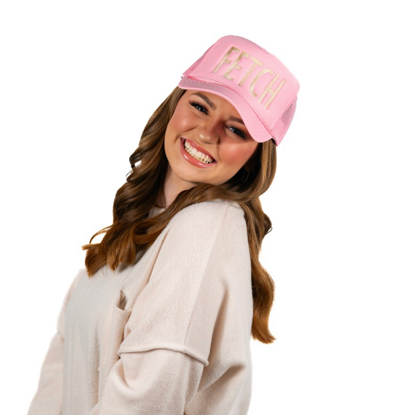 A woman beams while wearing the Fetch Pink Trucker Hat from Banks + Cove. The trendy hat with FETCH adds a splash of pink flair as her long, wavy brown hair flows over her light top, set against a simple white background.
