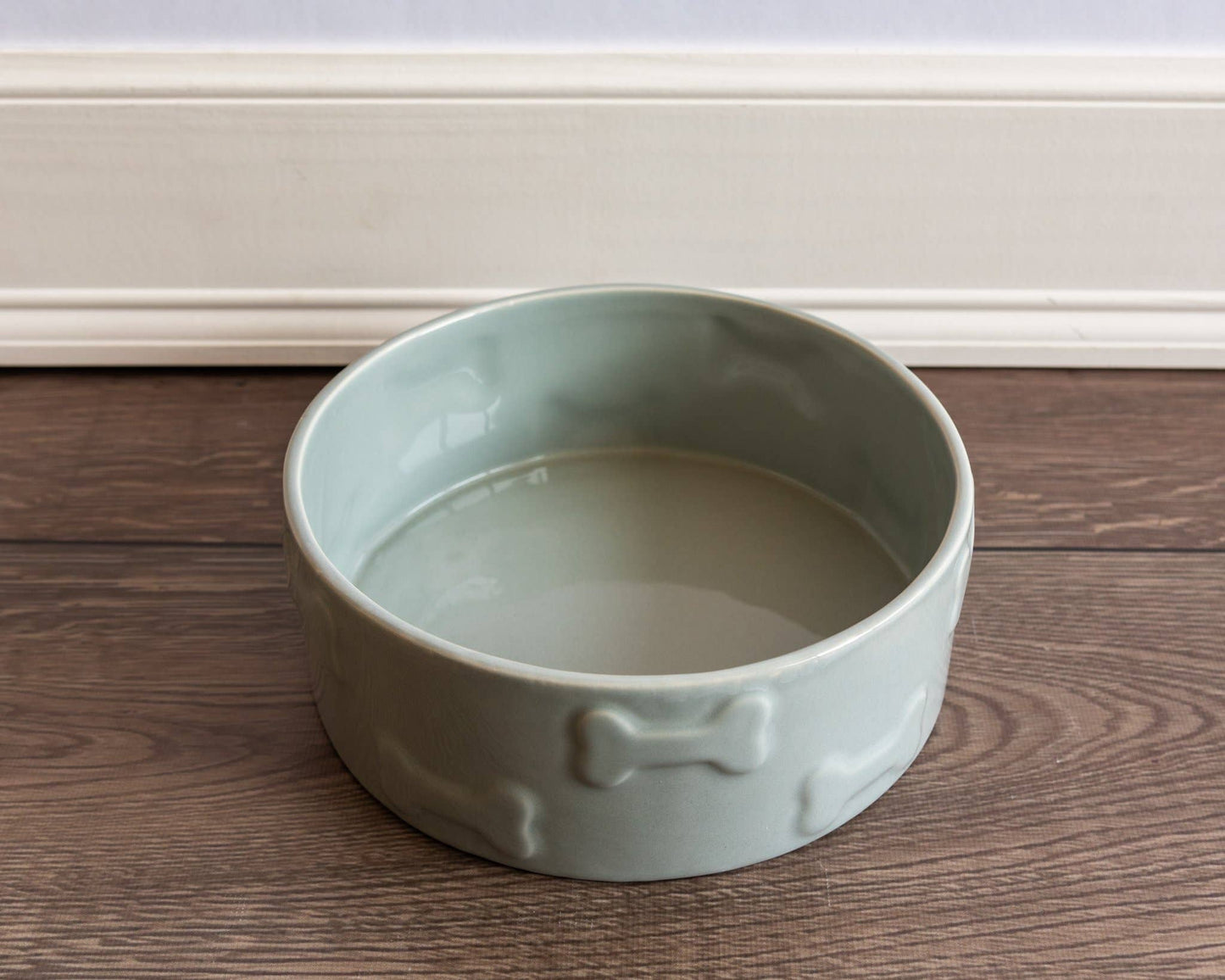A Manor Grey Pet Bowl by Park Life Designs, made from premium-quality ceramic with bone designs, sits elegantly on a wooden floor against a white wall.