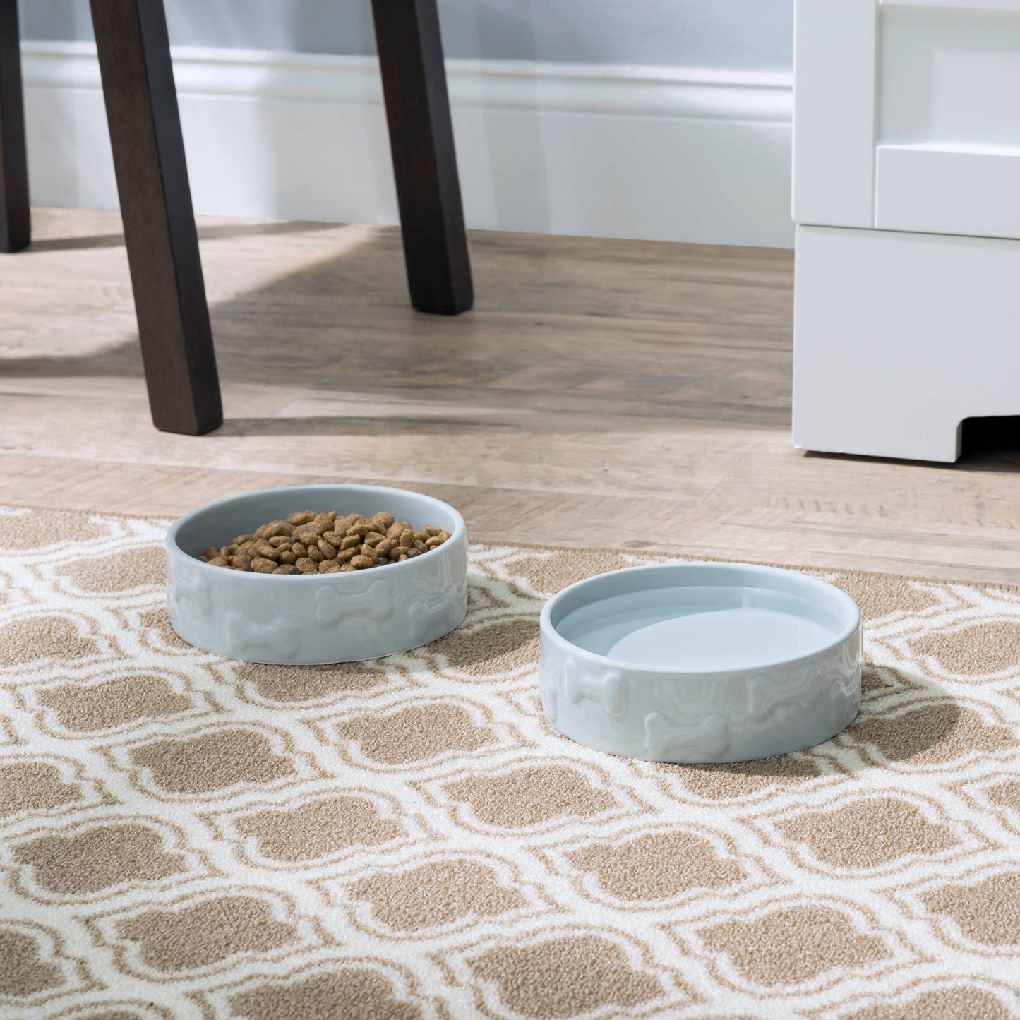 Two Manor Grey Pet Bowls from Park Life Designs sit on a patterned rug near a wooden chair and white cabinet. One ceramic bowl is filled with kibble, the other with water, all atop a wooden floor that complements the neutral-toned decor.