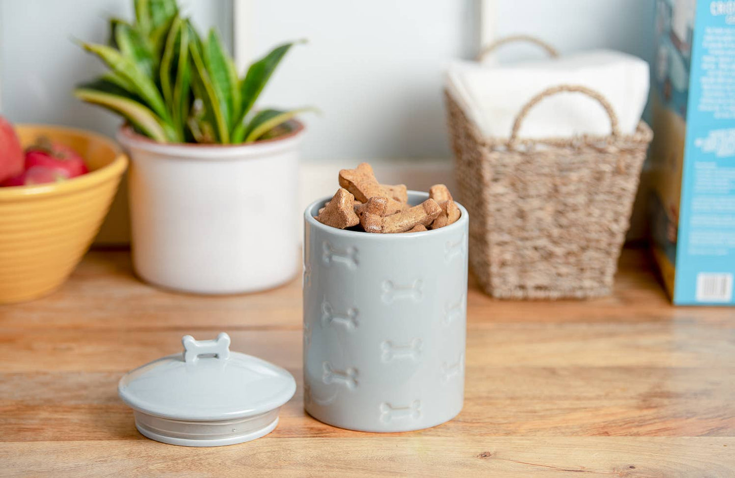A Park Life Designs Manor Grey Treat Jar with a bone pattern contains treats on a wooden countertop. Its airtight silicone-sealed lid is beside it, with a potted plant, dish basket, and apple bowl in the background—ideal for pet-friendly kitchens.