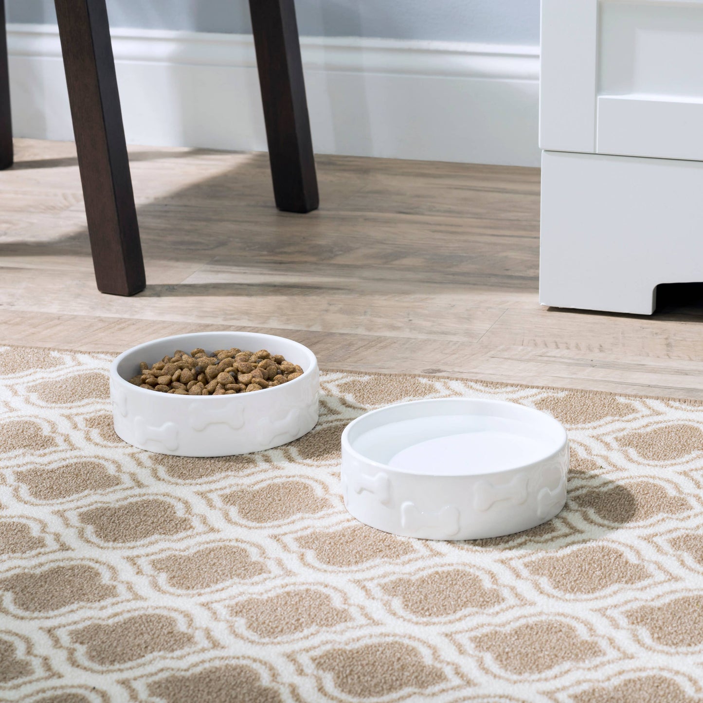 Two Manor White Pet Bowls by Park Life Designs sit on a patterned rug; one full of dry food, the other awaits use. Made from FDA-approved ceramic, they are dishwasher and microwave safe. On a light wood floor, near white furniture, they blend style with practicality effortlessly.
