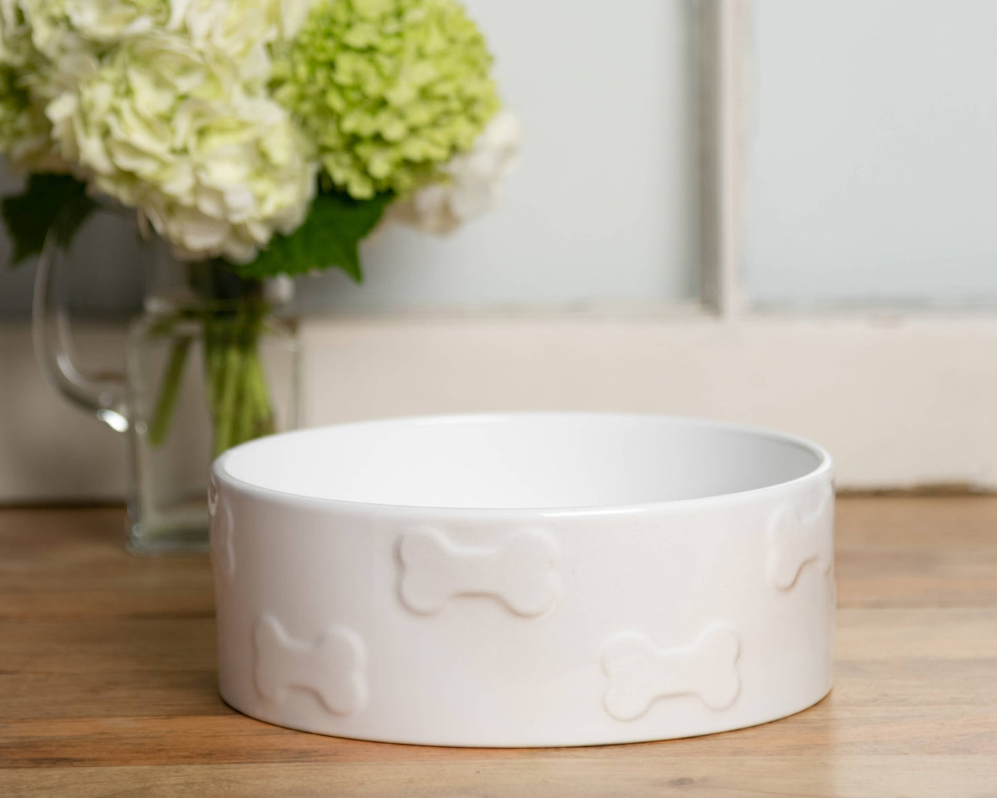 A Park Life Designs Manor White Pet Bowl, featuring bone motifs, sits on a wooden surface. Made of FDA-approved ceramic, its dishwasher and microwave safe. Behind it is a glass vase containing white and light green hydrangeas.