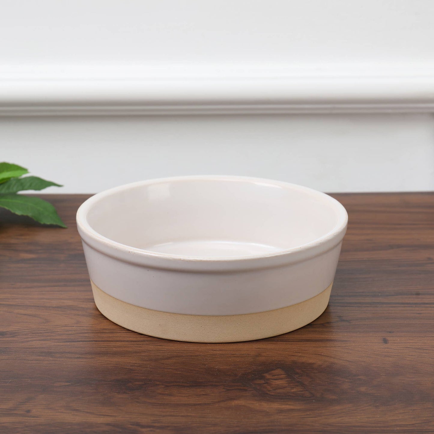 The Nordic Cream Pet Bowl by Park Life Designs, featuring a round ceramic design with a white top and potters clay base, exudes style on a wooden surface. A green plant is partially visible in the corner, set against a white background.