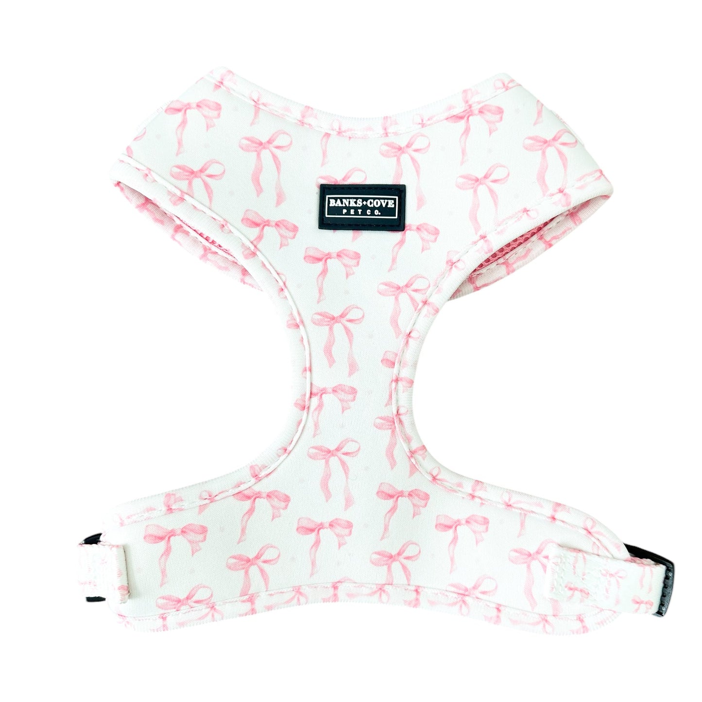 Introducing the Coquette Pooch- Dog Harness by Banks + Cove: a chic white harness with pink bows for stylish pets. It boasts a sleek black label, adjustable straps, and a side buckle for comfort and style.