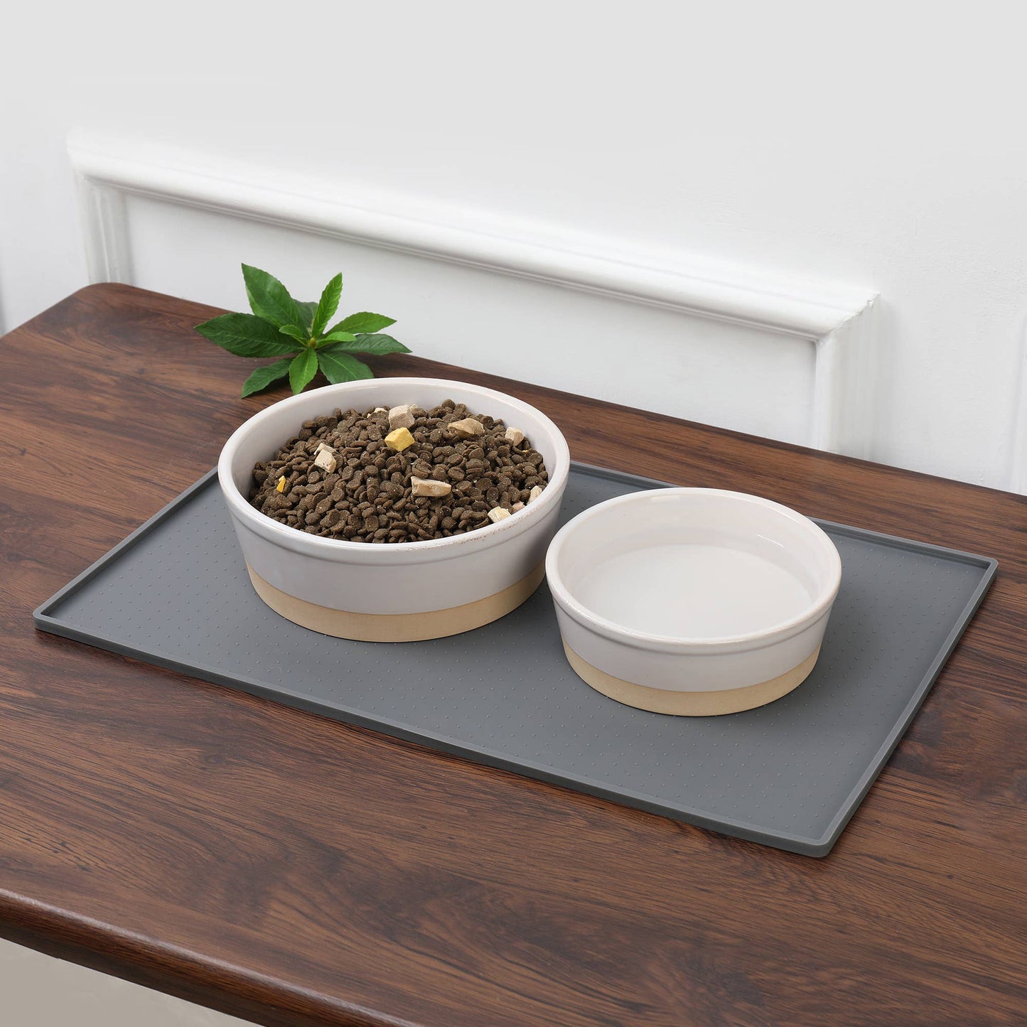 On a wooden table, two ceramic pet bowls are placed on a Park Life Designs Silicone Mess Free Pet Placemat. One bowl has dry pet food, the other water, and a small green plant adds nature in the backdrop against a white wall.