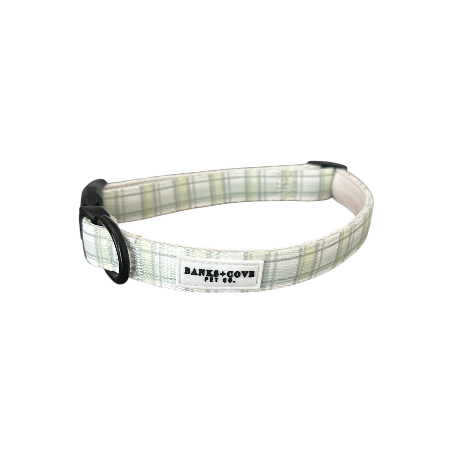 The Plaid About You Dog Collar from Banks + Cove is crafted using durable, lightweight materials in a light green and white plaid pattern. It features an adjustable fit, black buckle, D-ring, and a tag reading BANKS + COYE PET CO.