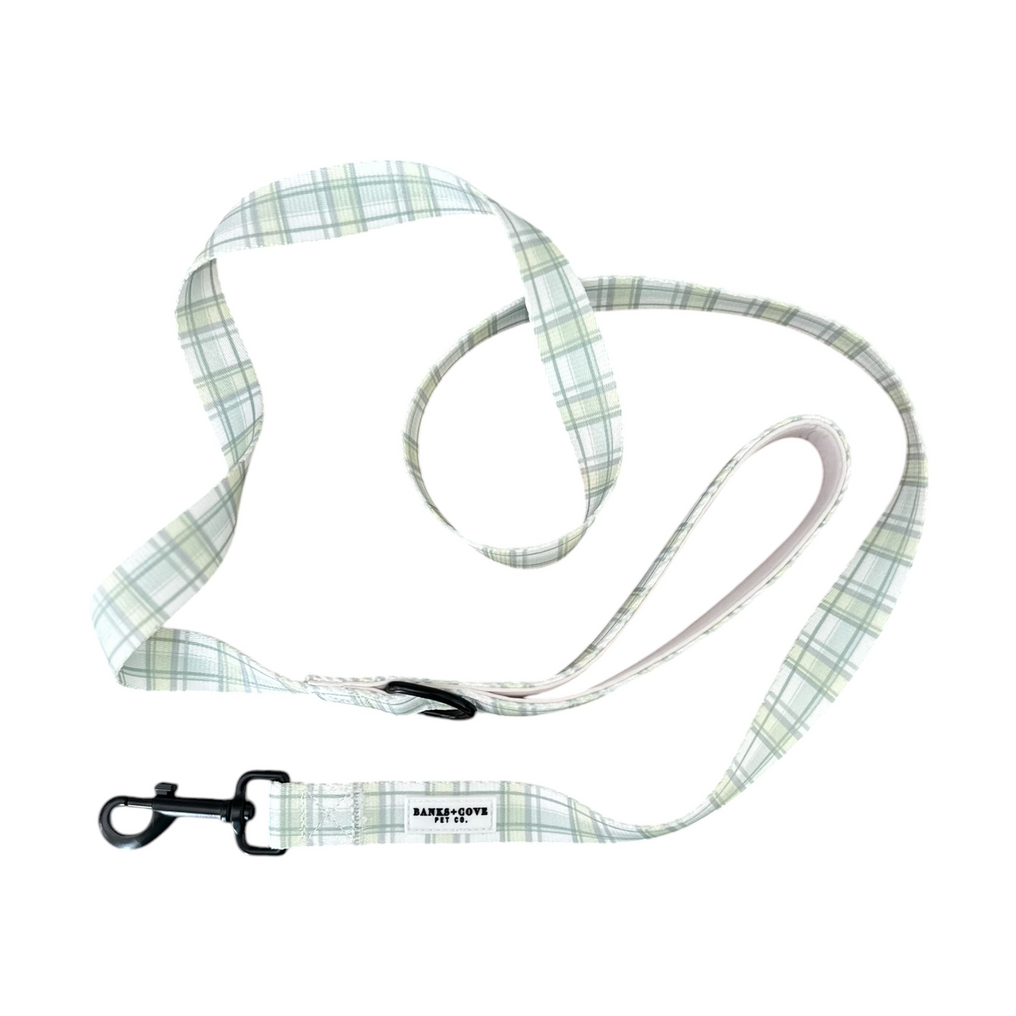 The Plaid About You- Dog Leash by Banks + Cove features a timeless light green plaid design, comfortable grip handle loop, and metal clip. Its subtle intersecting lines and small rectangular tag near the clip make it stylish and practical for daily walks.