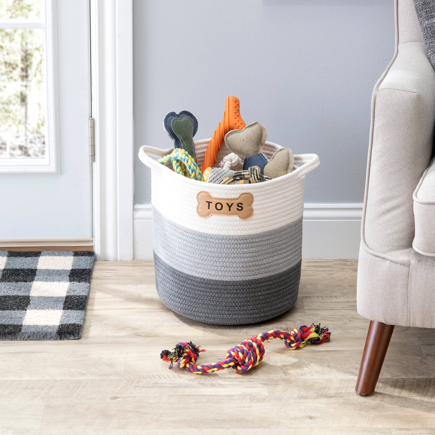 The Park Life Designs Livra Pet Toy Basket, in gray and white with a Toys label, provides a chic storage solution for bones and chew toys made of organic cotton. A colorful rope toy lays on the wooden floor next to the basket, near a gray chair and a plaid rug by the door.
