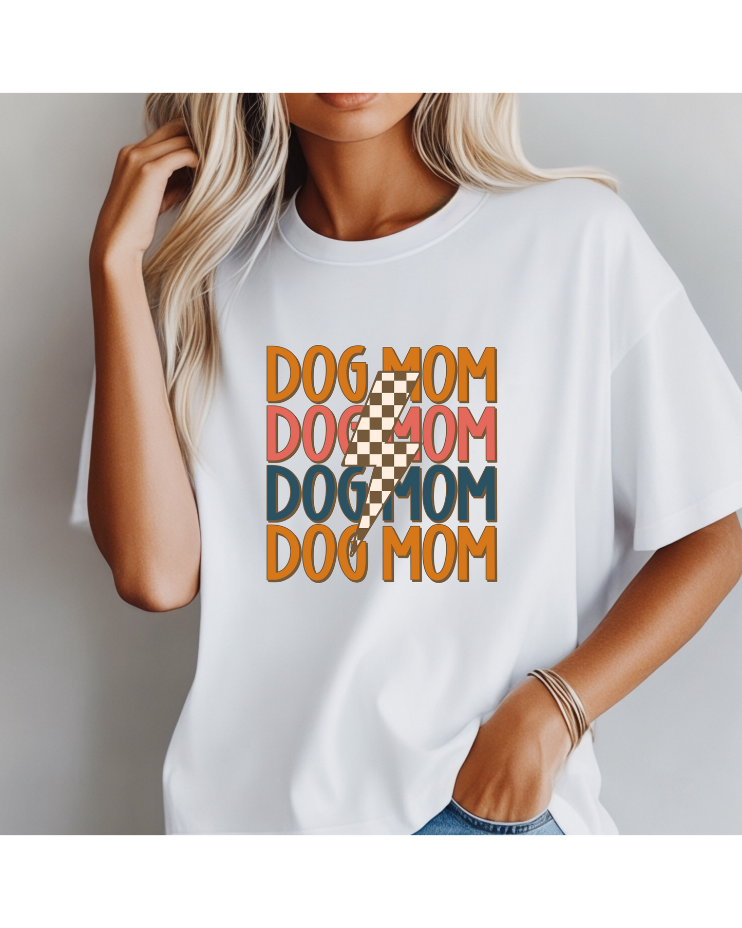 A woman with long blonde hair sports the Checkered Vibes Dog Mom Tee by Banks + Cove, featuring vibrant DOG MOM text. Exuding classic Dog Mom Energy, she casually holds her hair to one side while her other hand rests in her pocket.