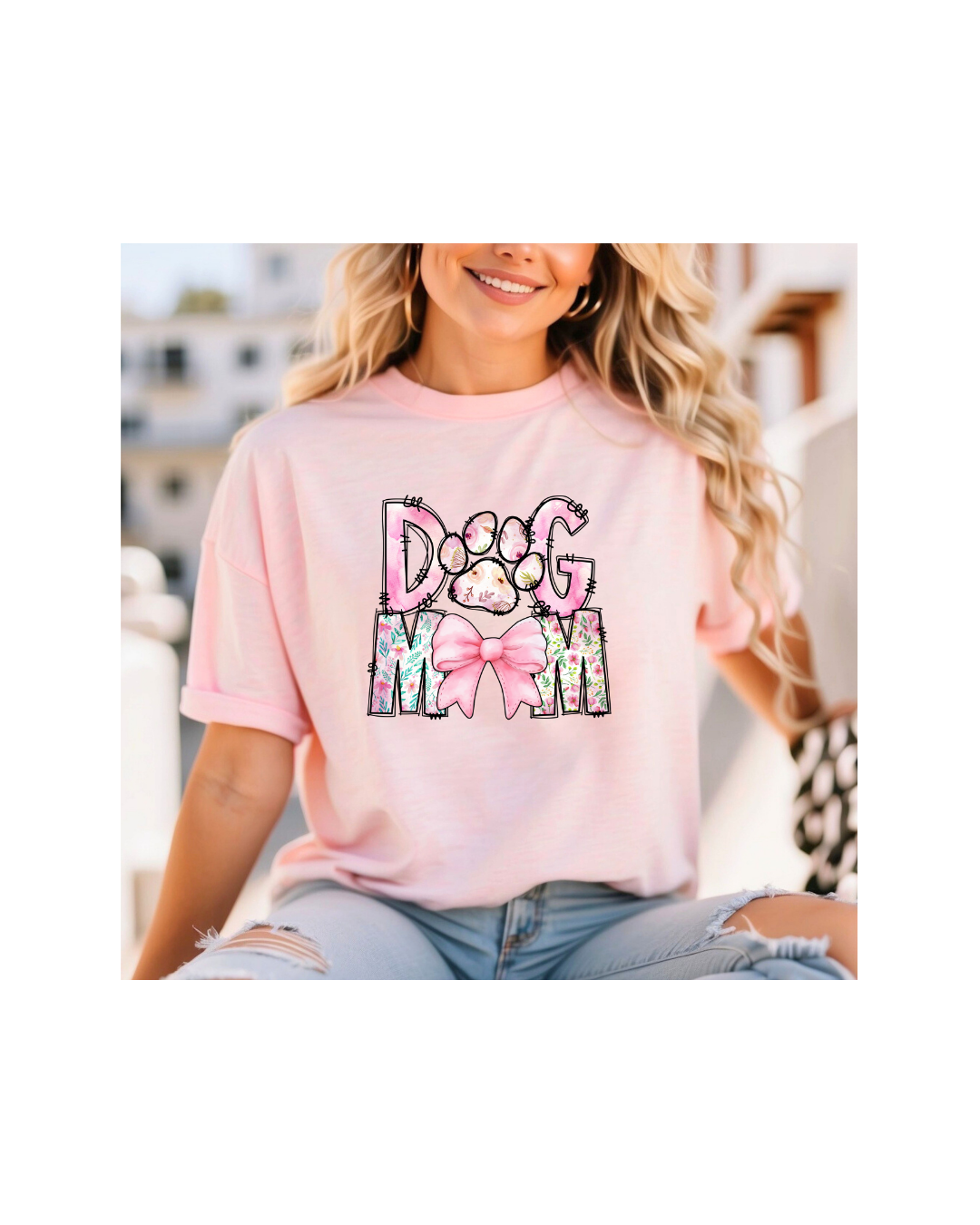 A person wearing a Paws & Petals Dog Mom T-Shirt by Banks + Cove, featuring watercolor florals and a pink bow, sits outdoors on a sunny day with long, wavy hair and smiles against a backdrop of buildings.