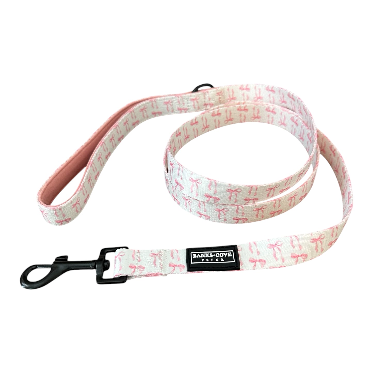 The Banks + Cove Coquette Pooch Dog Leash showcases a white design with pink bows and durable webbing lined with soft pink fabric. A robust black metal clasp provides secure attachment, while its padded handle ensures comfort on walks.