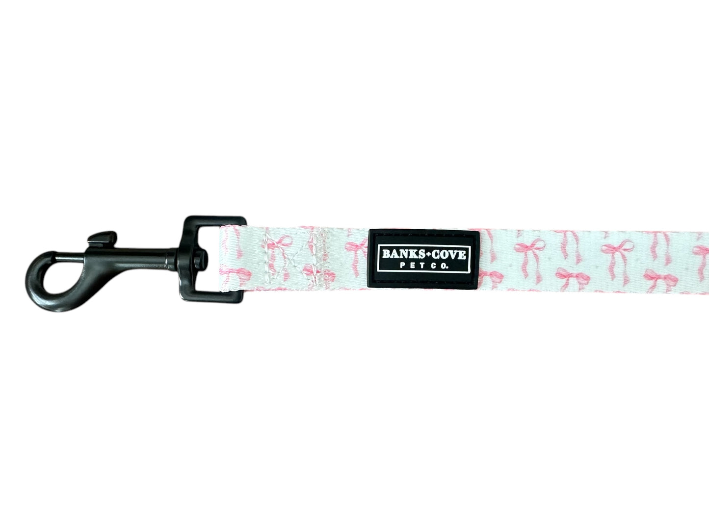 The Coquette Pooch Dog Leash by Banks + Cove features a white fabric strap with pink ribbon patterns and a black metal clip. Crafted with durable webbing, it includes a padded handle for comfort and bears a black Banks & Cove Pet Co. label near the clip.