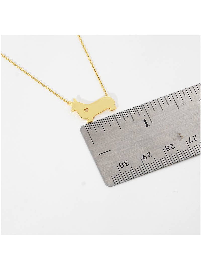 The Banks + Cove Corgi Love Necklace, featuring a gold-tone dog-shaped pendant about 1.5 cm long, is displayed beside a ruler with detailed centimeter and millimeter markers.