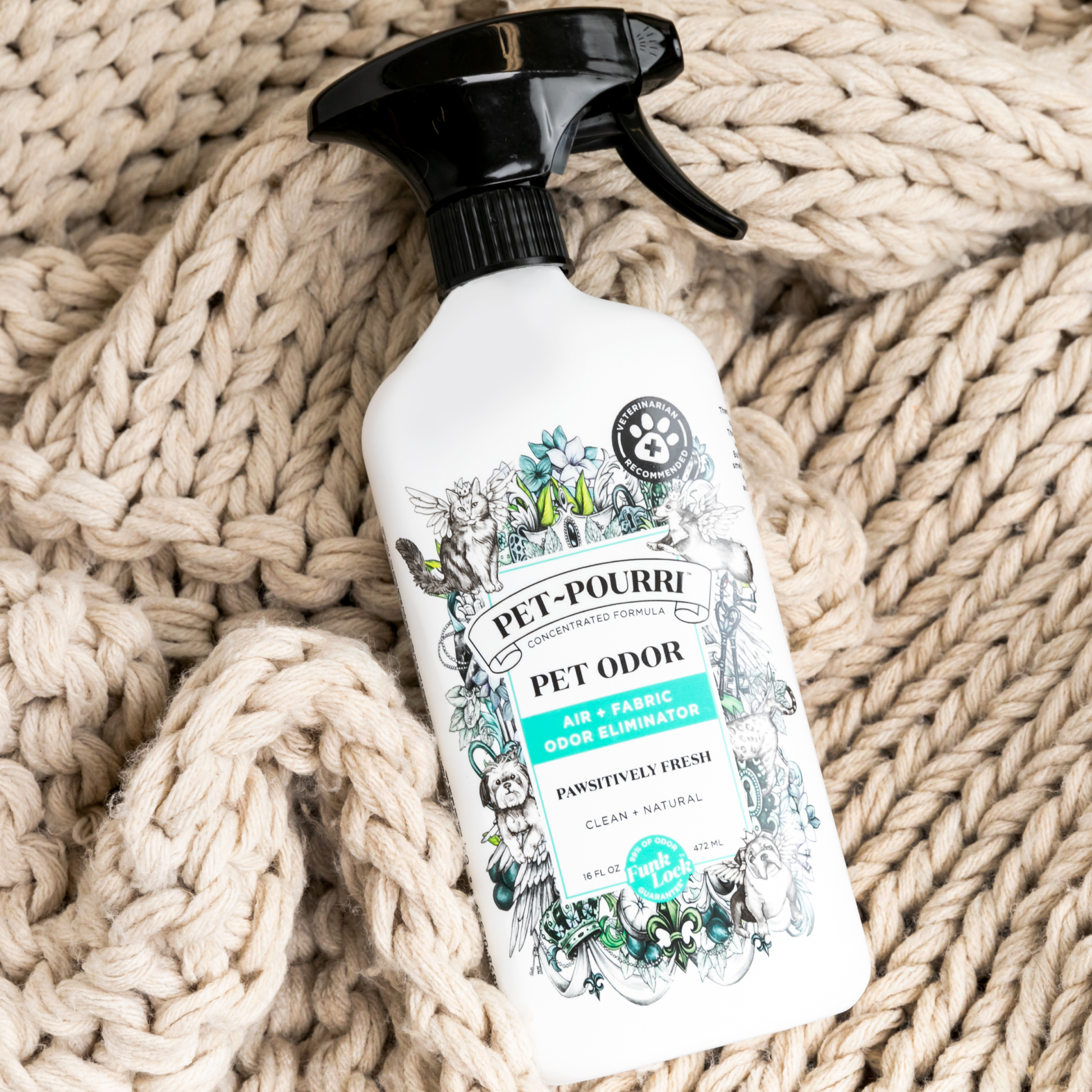 A bottle of Pet~Pourri Pawsitively Fresh Air+Fabric Odor Refresher 16oz sits on a knitted cream-colored blanket. The floral and paw print label complements the white spray bottle and black nozzle, featuring pet-friendly essential oils. Brand: ~Pourri.
