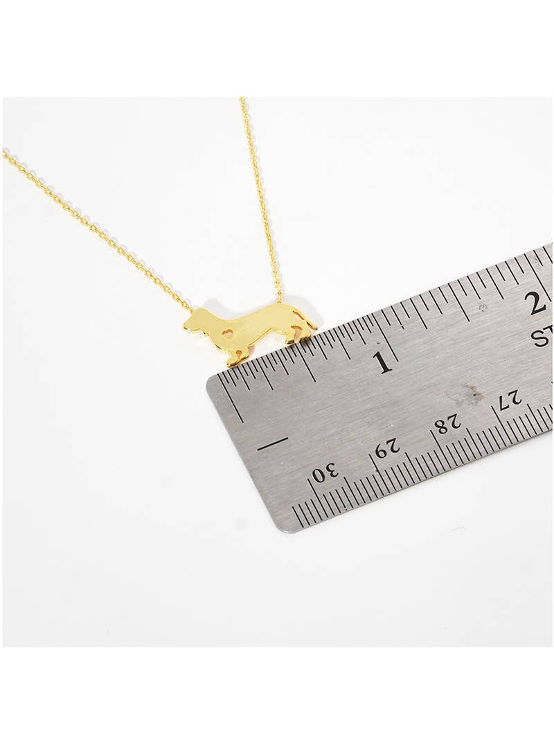 The Banks + Cove Dachshund Love Necklace features a charming gold-tone dog-shaped pendant, ideal for dog lovers. A silver ruler alongside it displays a length of about 2.5 cm, marked in black for precision.