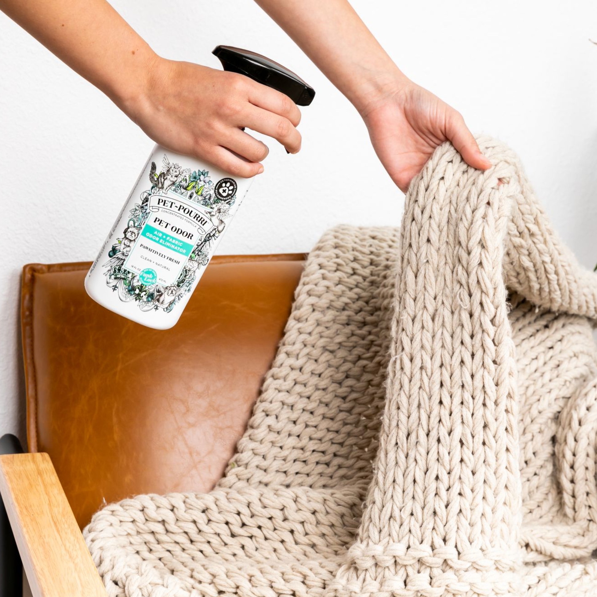 Someone sprays Pet~Pourri Pawsitively Fresh Air+Fabric Odor Refresher on a large, cream-colored knitted blanket over a brown leather chair. The bottle, by ~Pourri, is decorated with floral designs and a label reading Pawsitively Fresh.