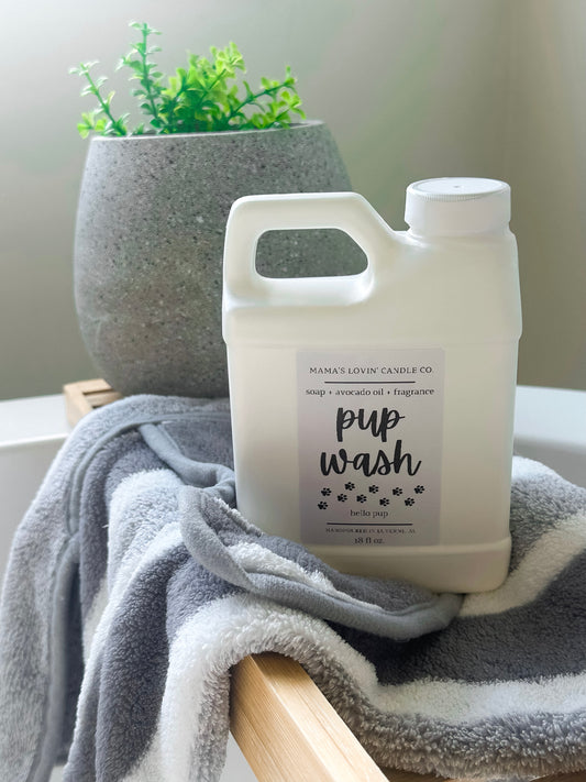 A white bottle labeled Mamas Lovin Pup Wash from Mamas Lovin sits on a striped towel atop a wooden bath tray, with a potted plant in the background, promising a long-lasting scent for your pup.