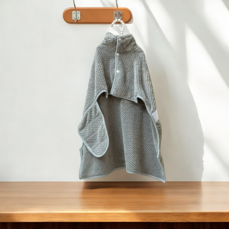 A Banks + Cove Bathrobe Drying Towel, soft and gray with a fluffy collar, hangs on a wooden hook, resembling an elegant poncho. Its shadow falls against the white wall, while a wooden surface adds to the minimalist decor, creating cozy dog bath time vibes.