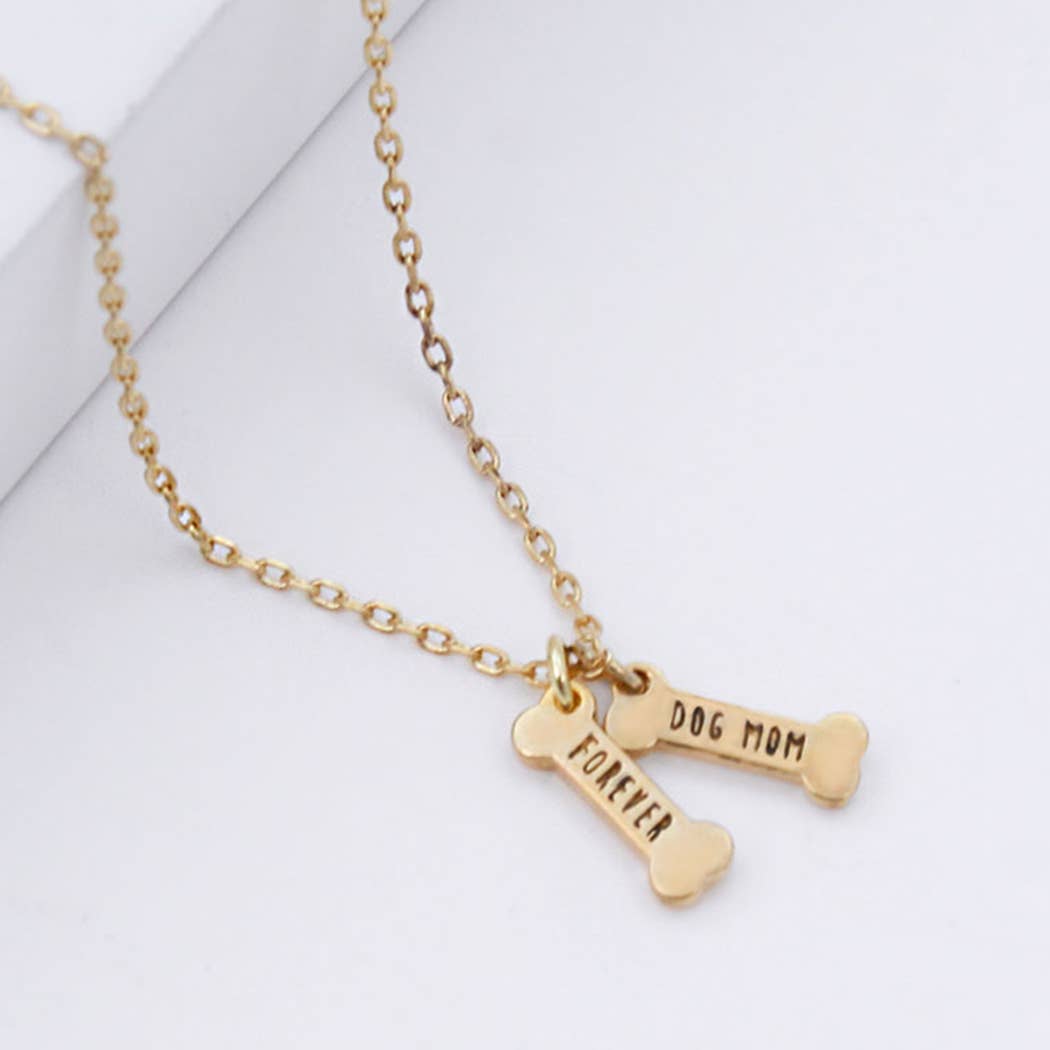 The Banks + Cove I Chews You – Dog Mom Bone Charm Necklace features a gold-dipped design with two bone-shaped pendants, one inscribed with FOREVER and the other with DOG MOM, elegantly displayed on a white surface.