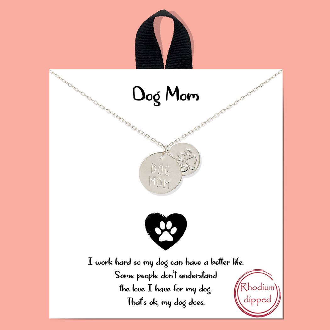 A Banks + Cove Gold-Dipped Dog Mom charm necklace with a paw print pendant sits on a display card that says, I work hard so my dog can have a better life. Some people dont understand the love I have for my dog—thats okay, my dog does. A Rhodium-dipped label is visible.