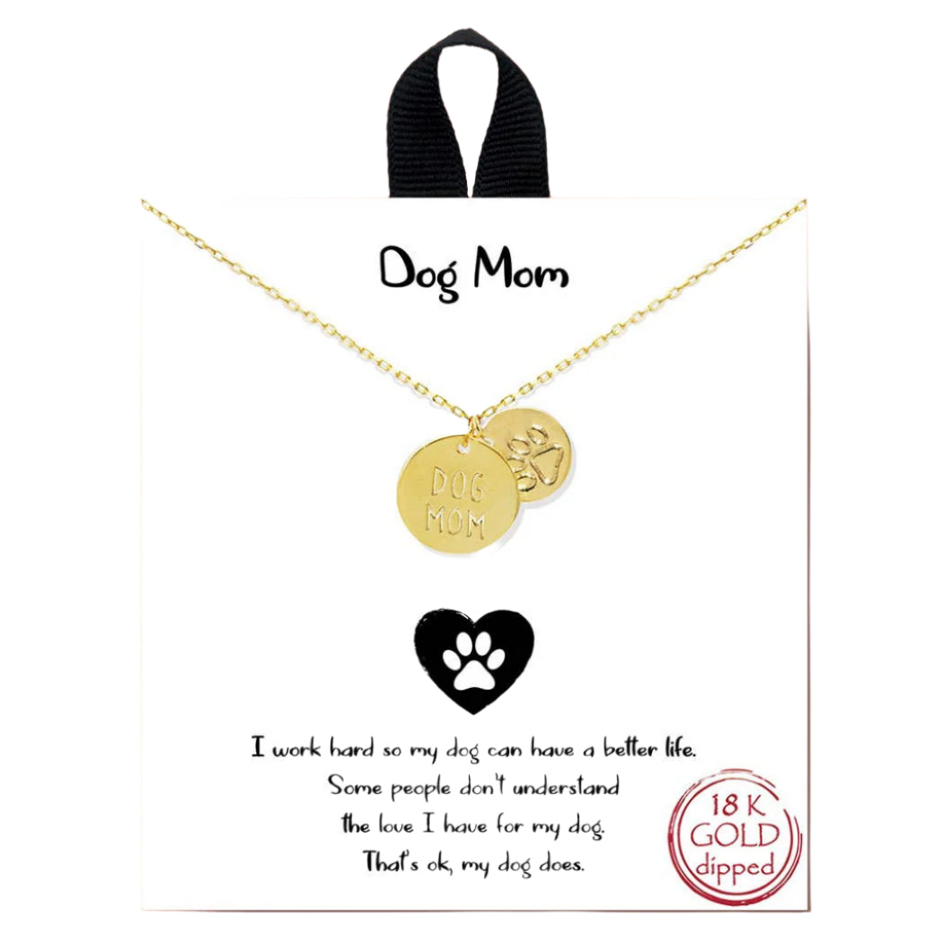 The Banks + Cove Dog Mom Charm Necklace features two pendants—one inscribed with Dog Mom and the other a paw print. It is 18K gold dipped, set against a pink background with text about love for dogs.
